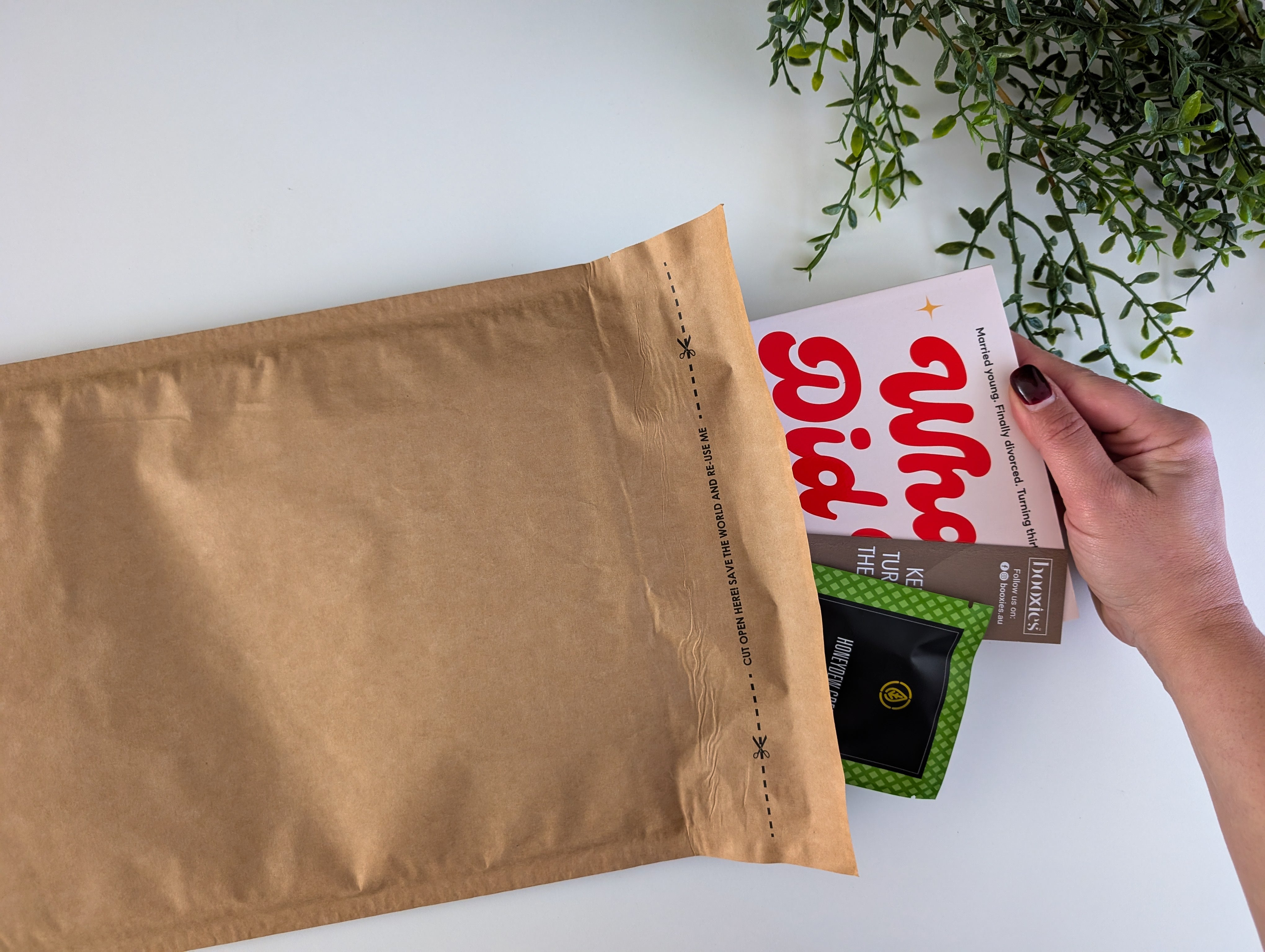 Person holding a box with 'What did i miss? book next to a brown envelope on a white surface with greenery. Aug 2025 boxless mystery booxies