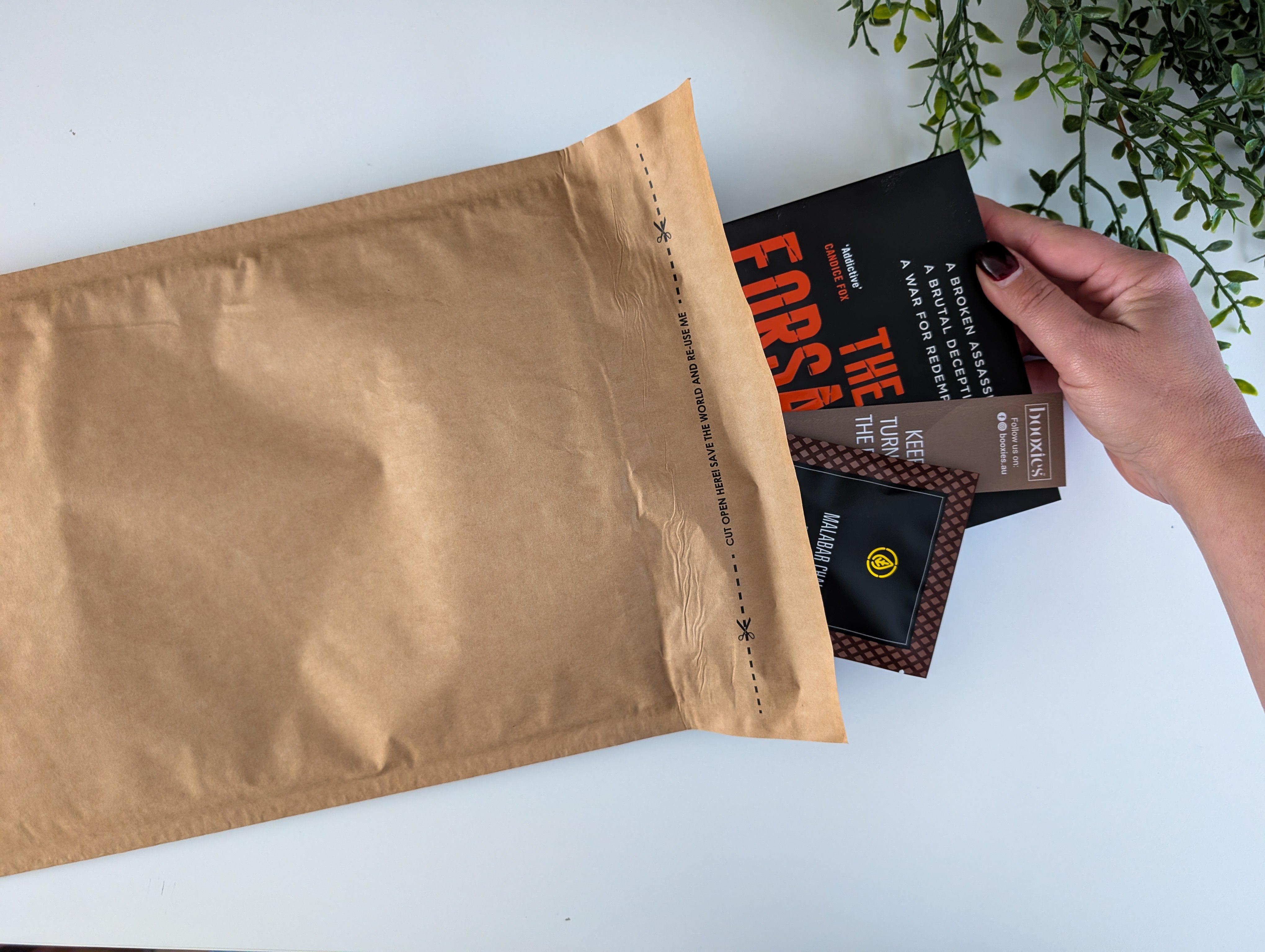 Person placing books into a brown envelope on a white surface with greenery in the background. this is Aug 2025 boxless mystery booxies