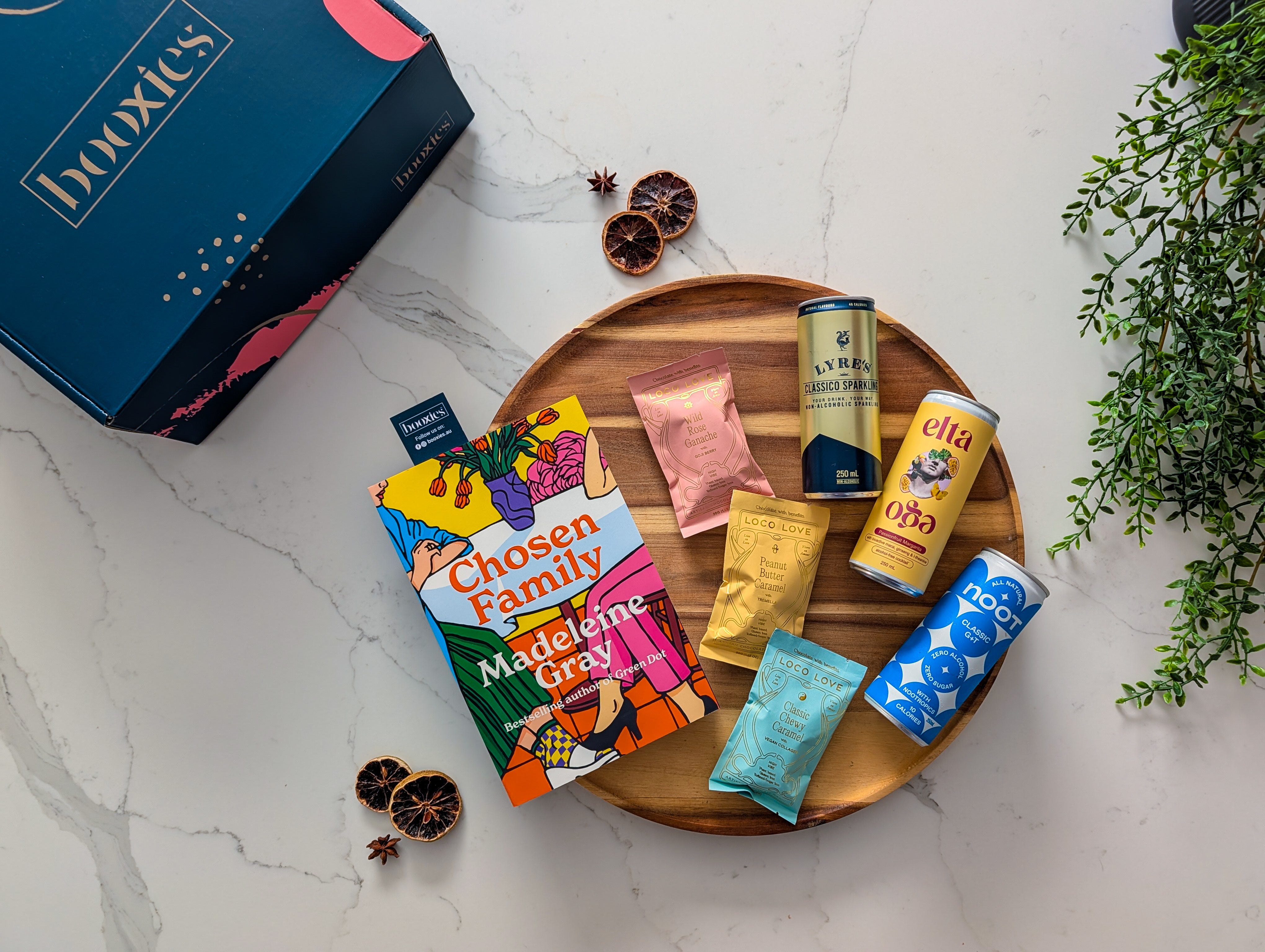 Assorted loco love chocolate and non-alcoholic drinks on a wooden tray with a 'booxies' branded box and a new book in the background. making up the Zero Edit curated booxies