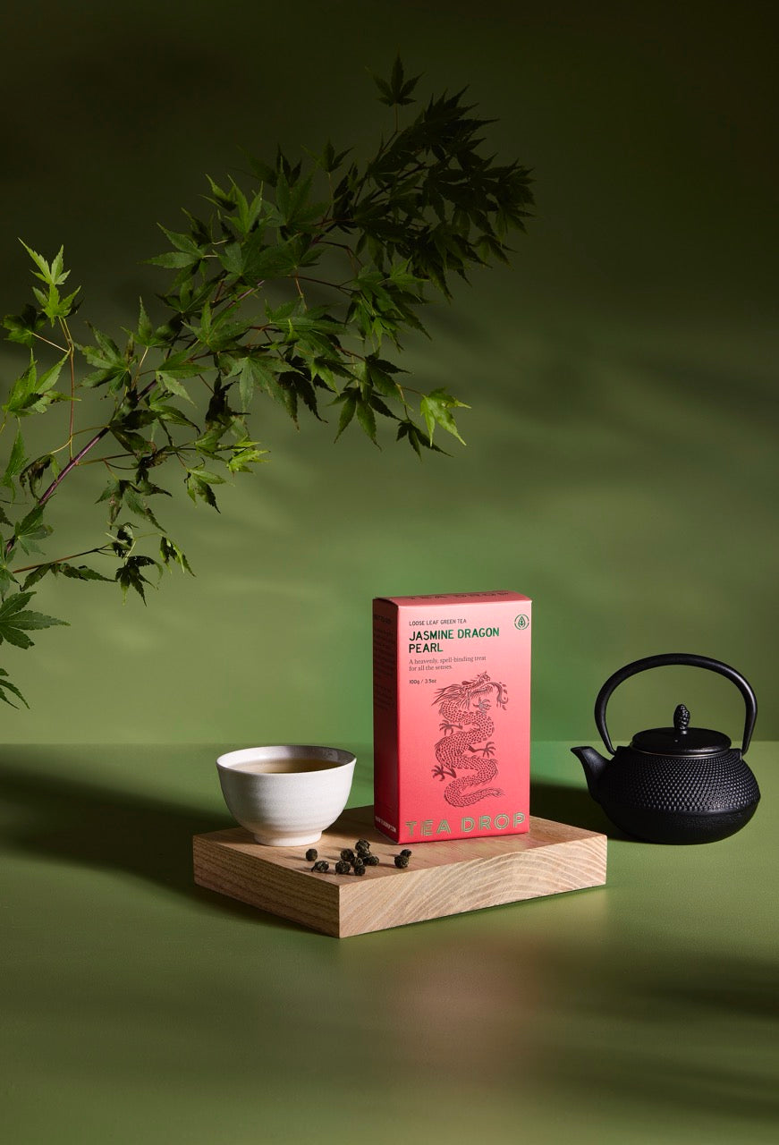 Tea packaging with a teacup, black teapot, and green leaves on a wooden platform against a green background