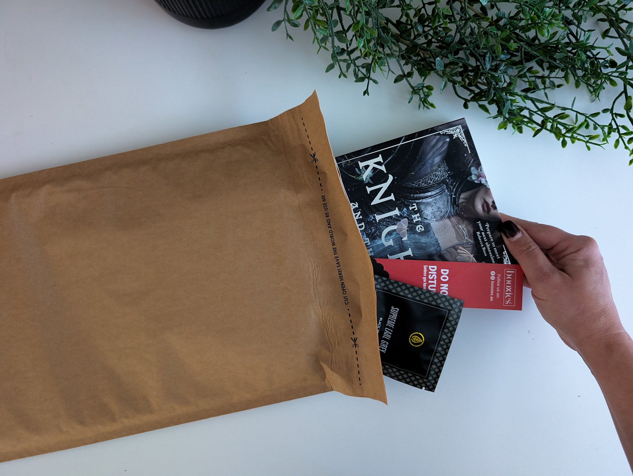 kraft envelope on a white surface with a hand holding a red bookmark, a tea pyramid and a book from our July Boxless Mystery Booxies.